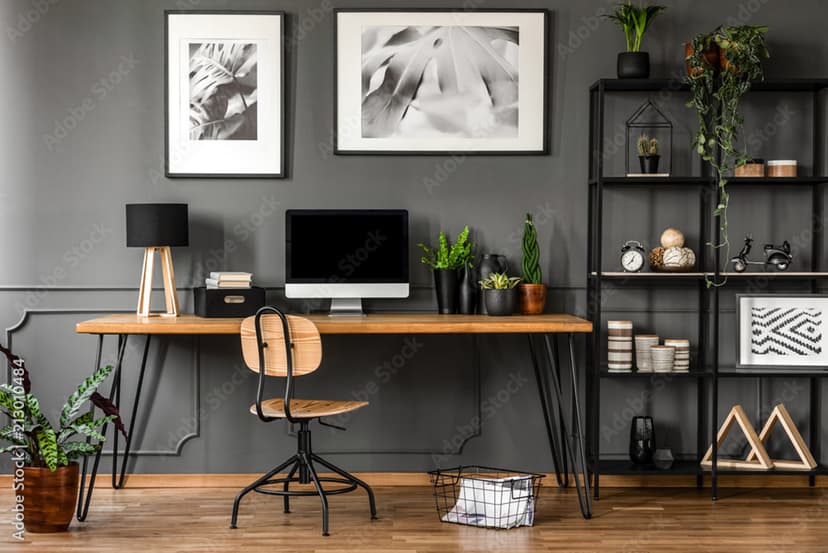 Modern flex room with a minimalist home office setup featuring a long wooden desk, black metal legs, computer monitor, indoor plants, black shelving unit with decor, and wooden flooring against a dark gray wall.