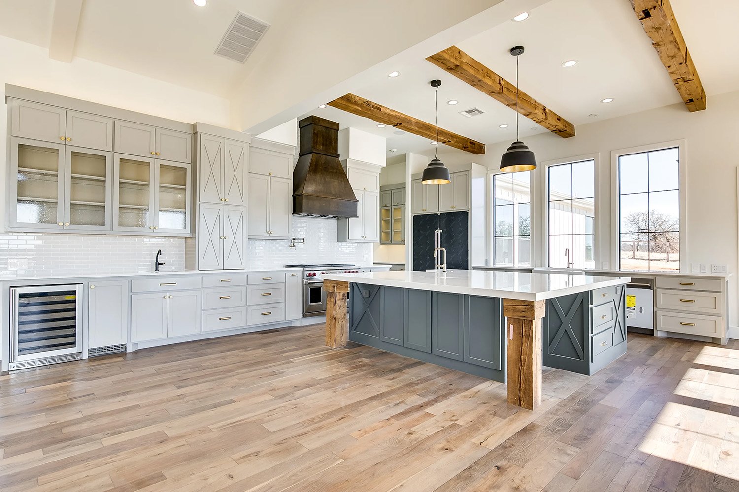 Spacious custom kitchen in Edmond, Oklahoma featuring exposed wood ceiling beams, a large center island with rustic wood accents, modern pendant lighting, light gray cabinetry, and expansive windows overlooking the outdoors.