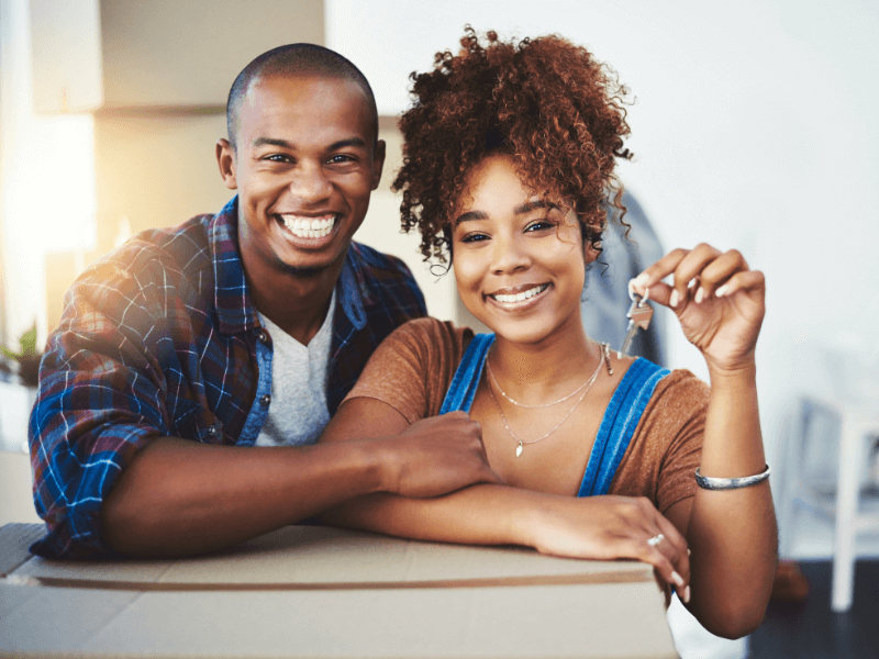Two smiling homeowners with a cardboard box and keys in hand