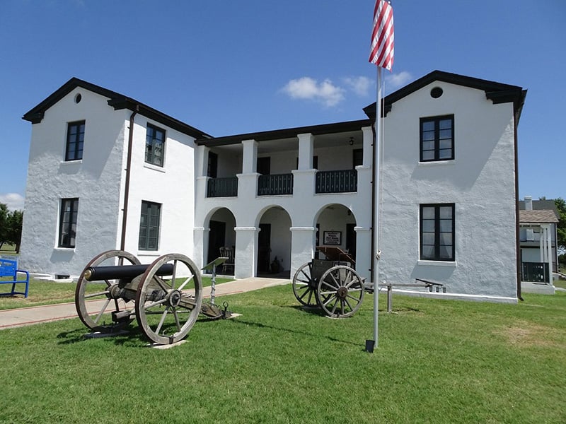 Historic Fort Reno in El Reno, Oklahoma, featuring a white two-story building with arched entryways, cannons on the lawn, and an American flag flying in front.