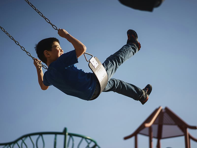 Child in a blue shirt enjoying an outdoor swing set under a clear sky.